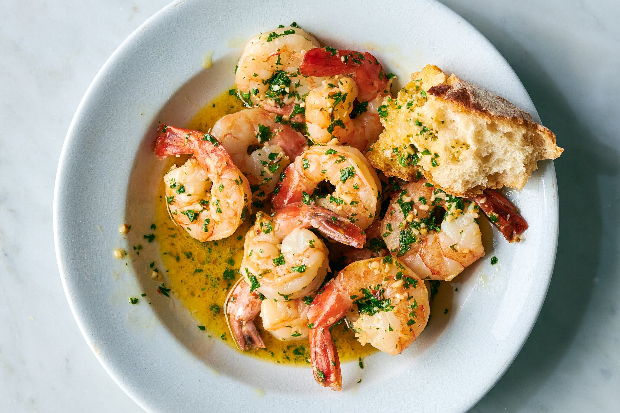 A white dinner plate is filled with parsley- and garlic-flecked shrimp and a hunk of baguette.