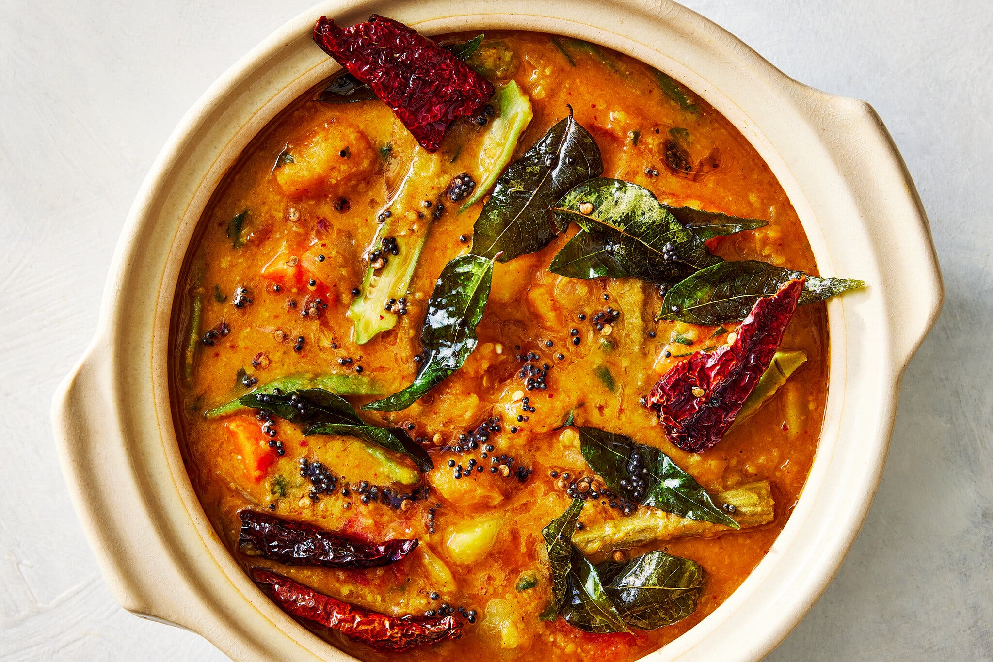 An overhead image of a red-orange stew topped with curry leaves and chiles.