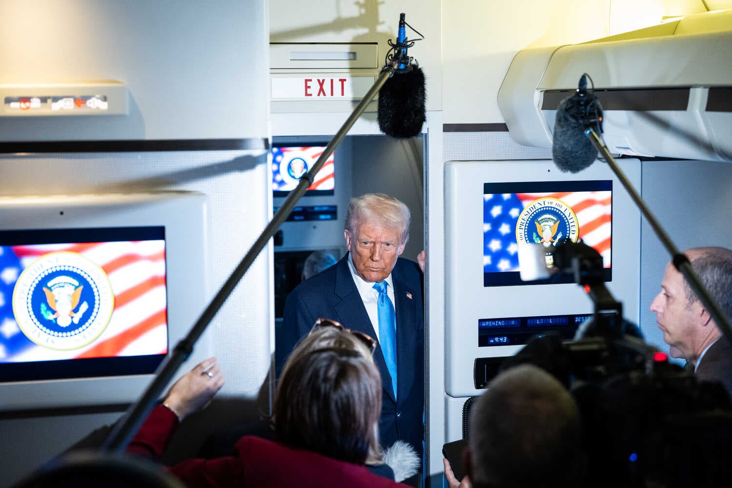A man in a dark suit talking with journalists on an airplane.