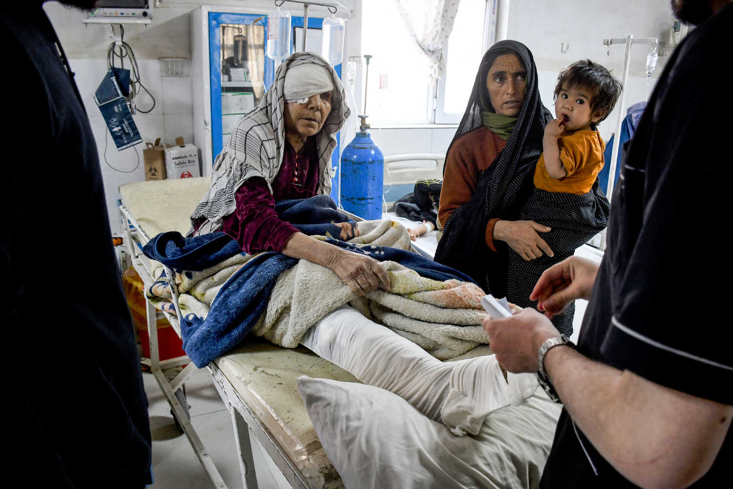 A woman in layers of clothes and a bandage covering her right eye sits up on a stretcher, next to another woman holding a baby.