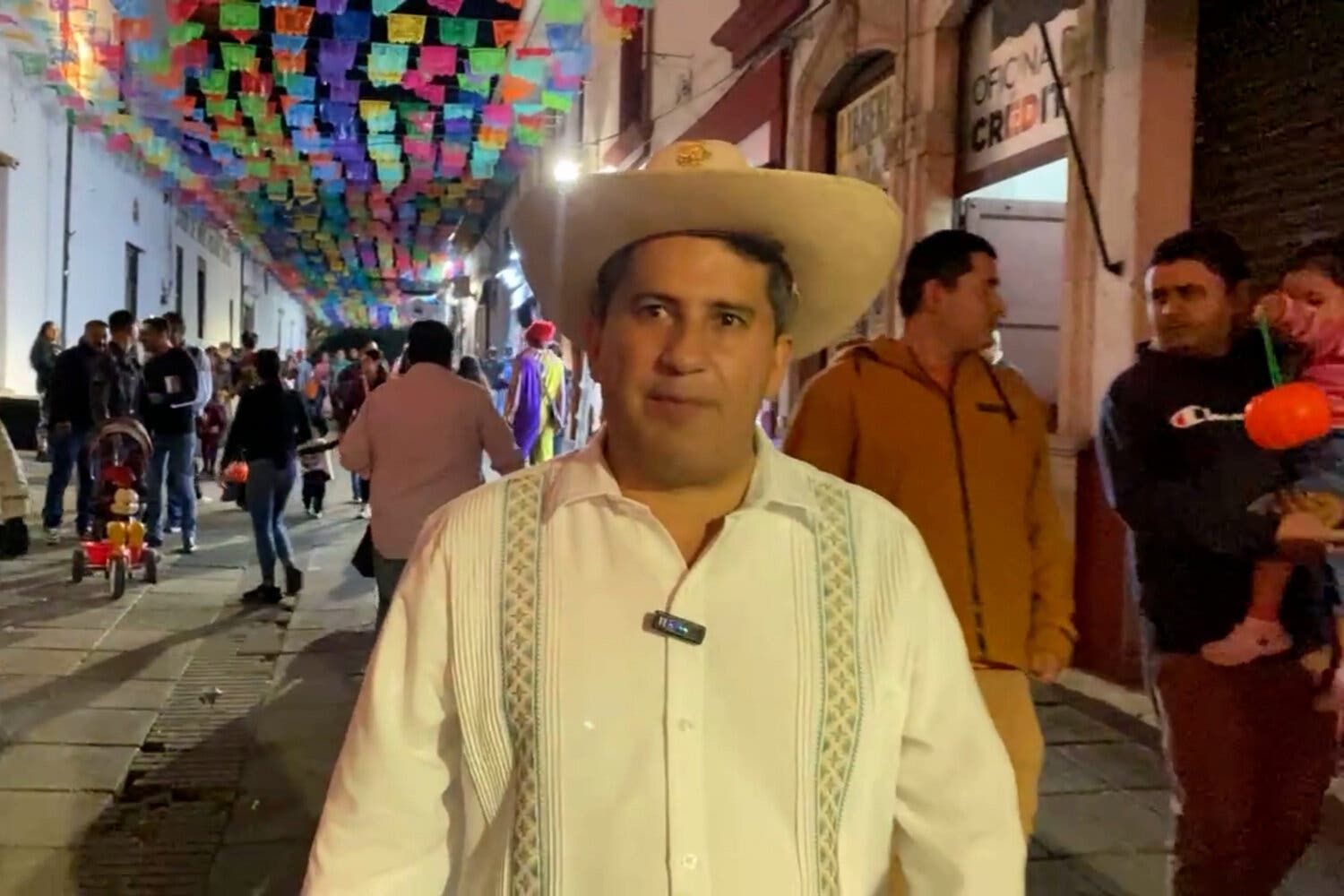 A man in a hat stands in an alley as police offers and others walk underneath paper decorations.