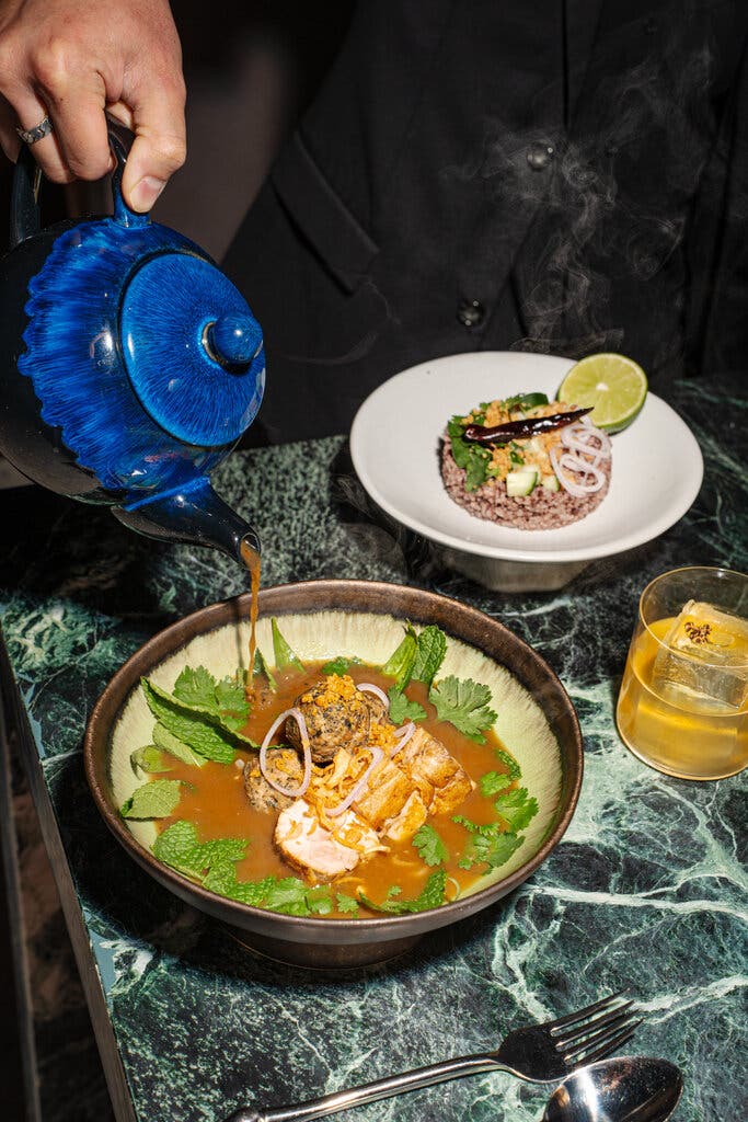 A person pours a broth into a bowl filled with chicken and herbs. A cocktail sits nearby.