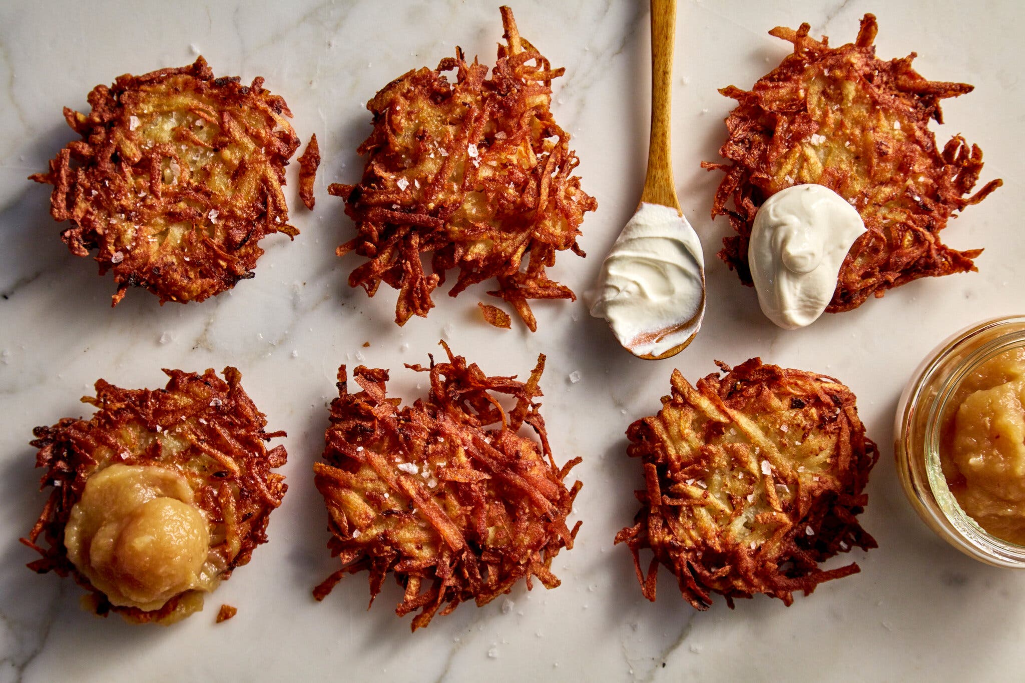 A marble surface with six latkes, some topped with a dollop of applesauce or sour cream.