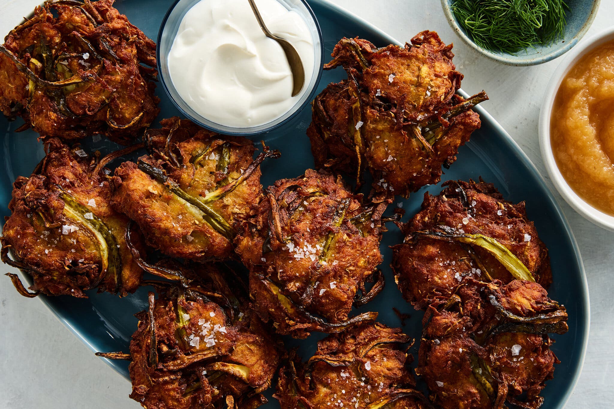 A batch of pickle latkes on a dark blue platter with sour cream, dill and applesauce.
