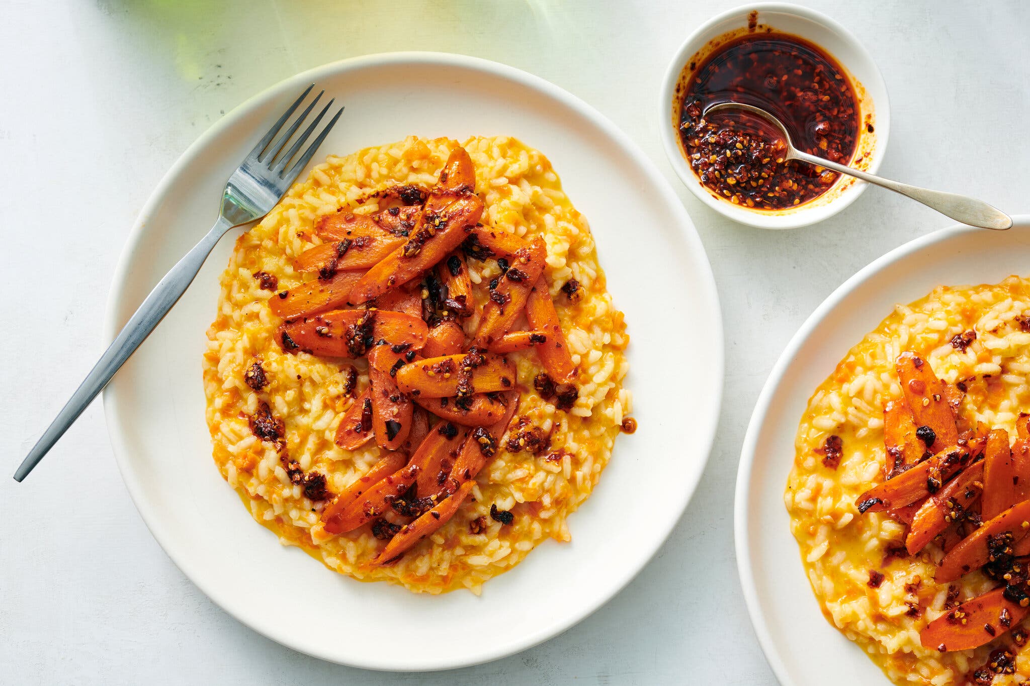 Two plates filled with carrot risotto and topped with cooked carrots sit next to a small bowl of chile crisp.