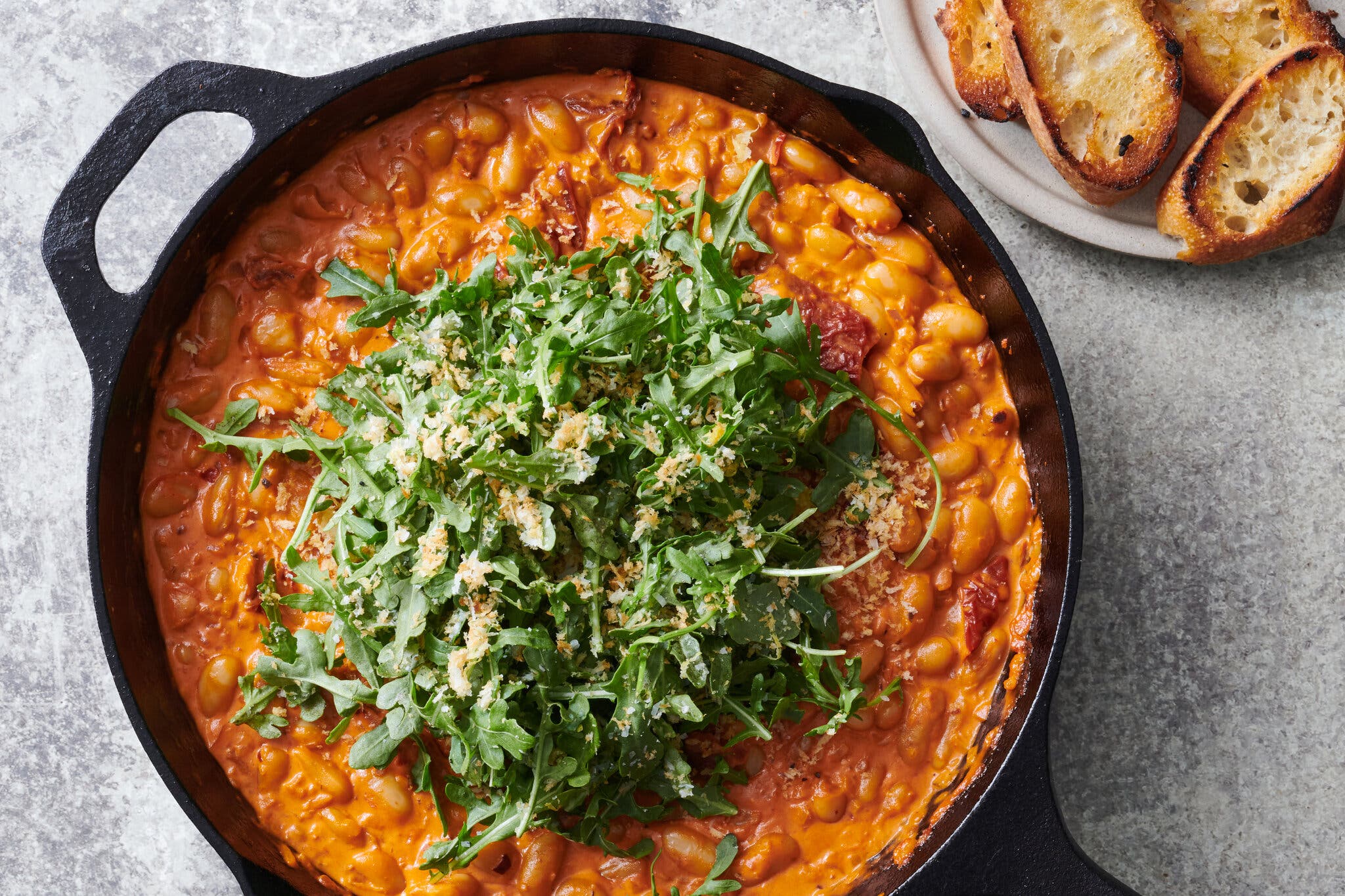 A large skillet is filled with beans in a red-orange sauce and topped with arugula and bread crumbs. A plate with toasted bread is also visible.