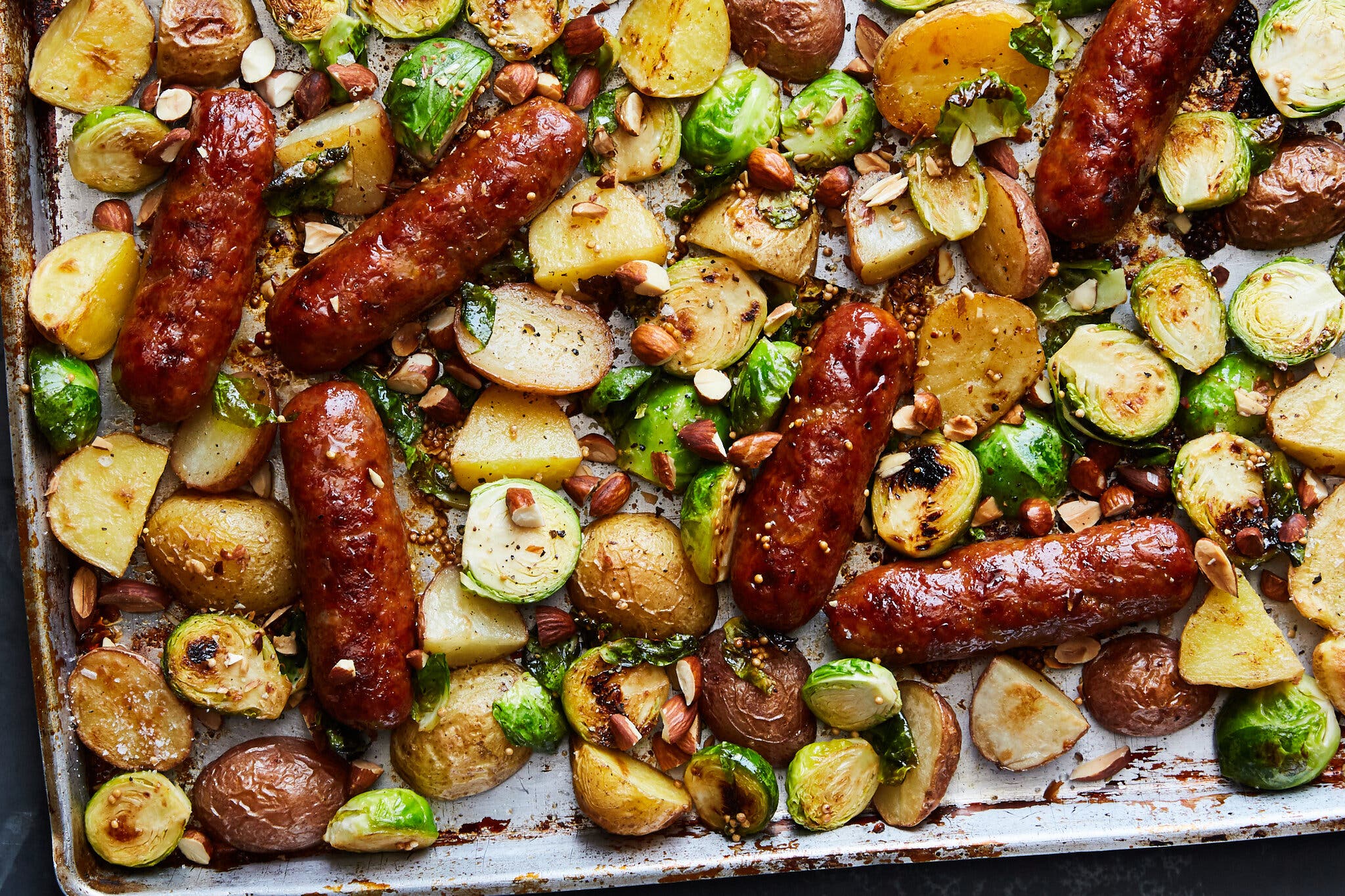 Roasted sausages, potatoes and brussels sprouts are spread out on a sheet pan.
