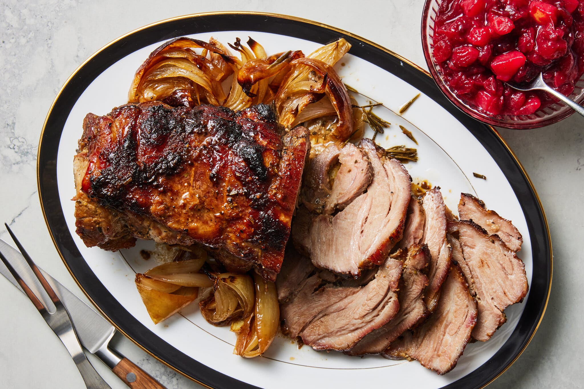 A pork roast surrounded by cooked onions sits on a platter next to a bowl of cranberry sauce.