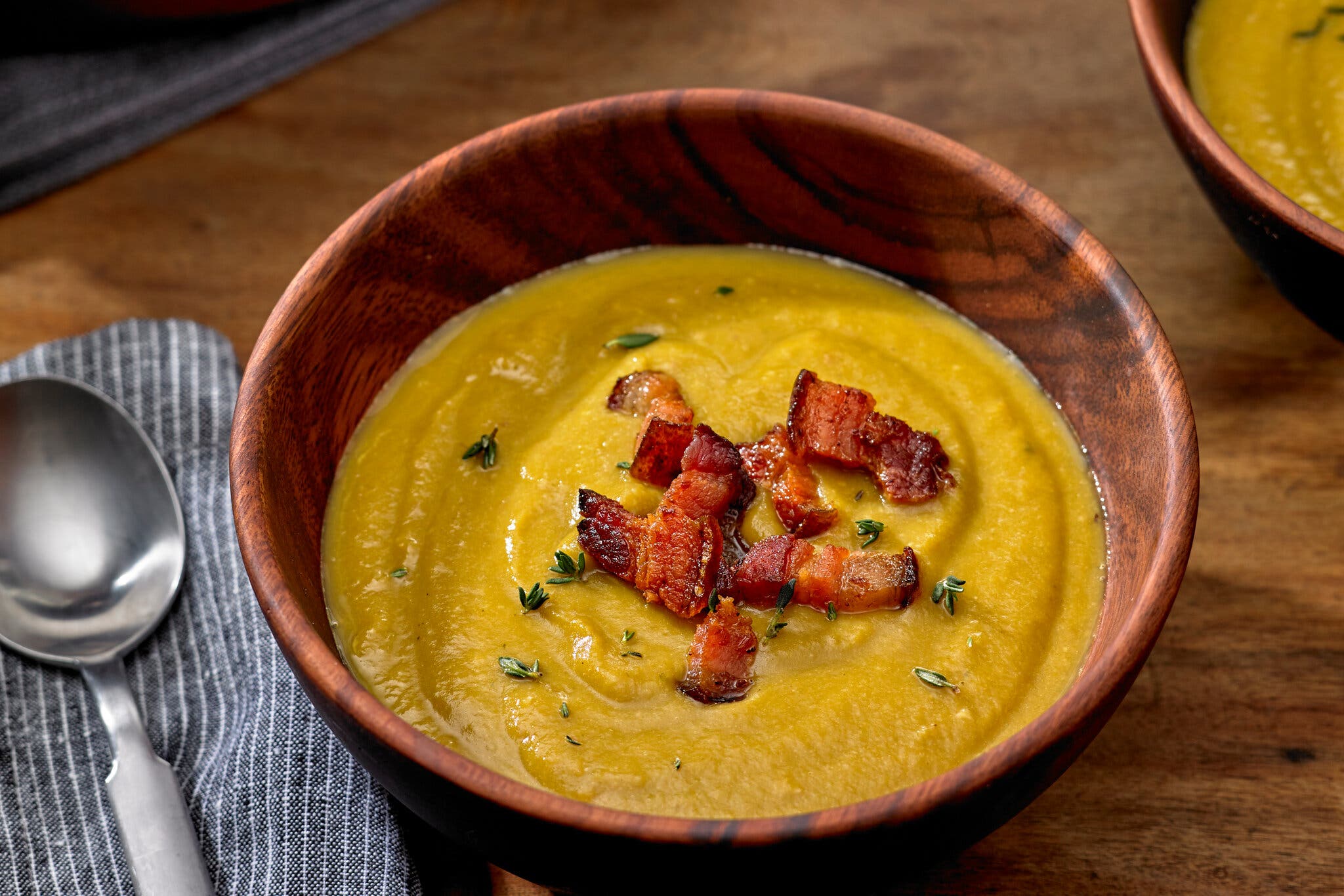 A wooden bowl filled with green-yellow split pea soup and topped with chunks of bacon sits next to a metal spoon on a striped napkin.