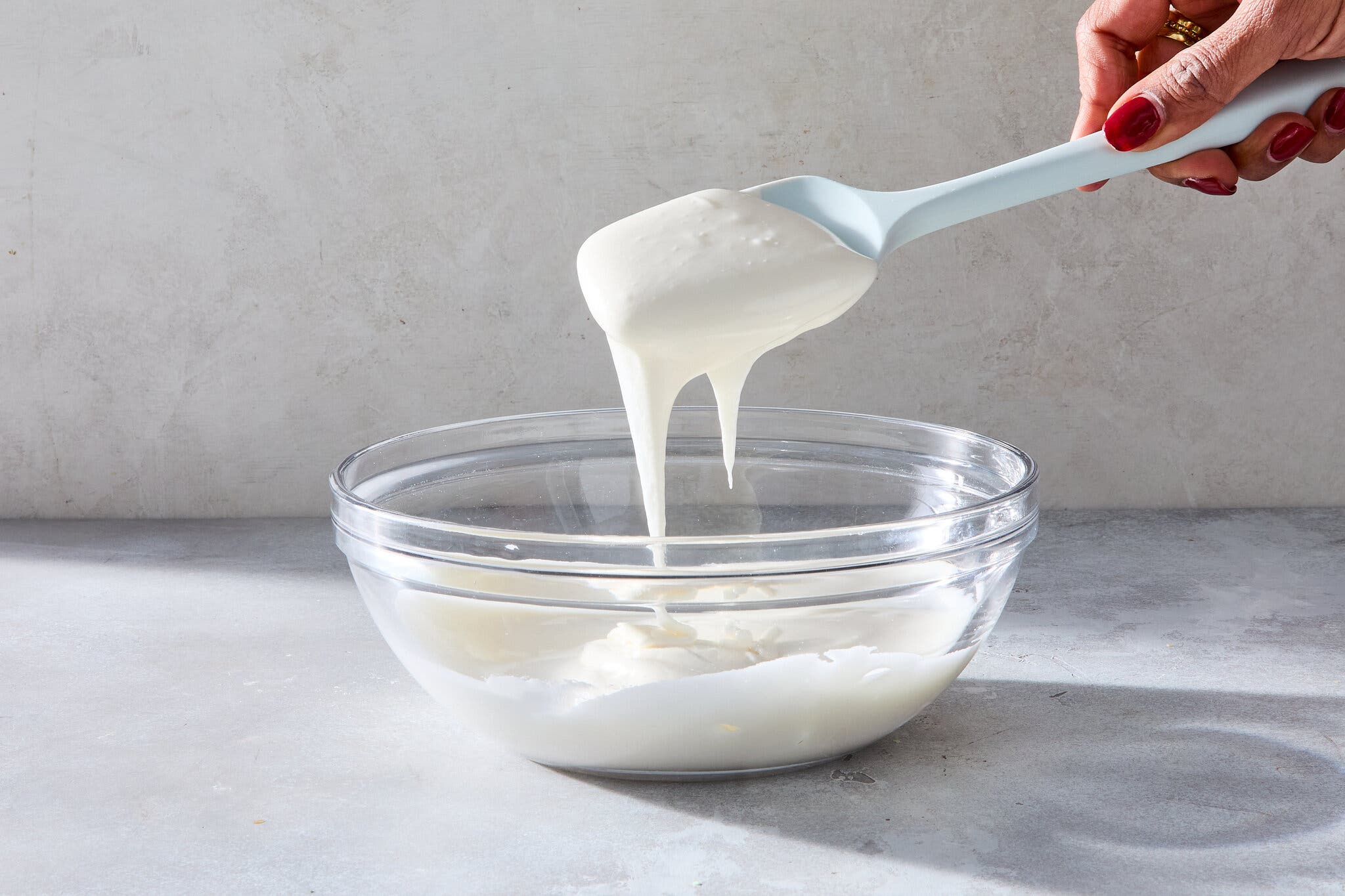 A side image of a clear bowl filled with icing. A hand holding a spatula is just above.