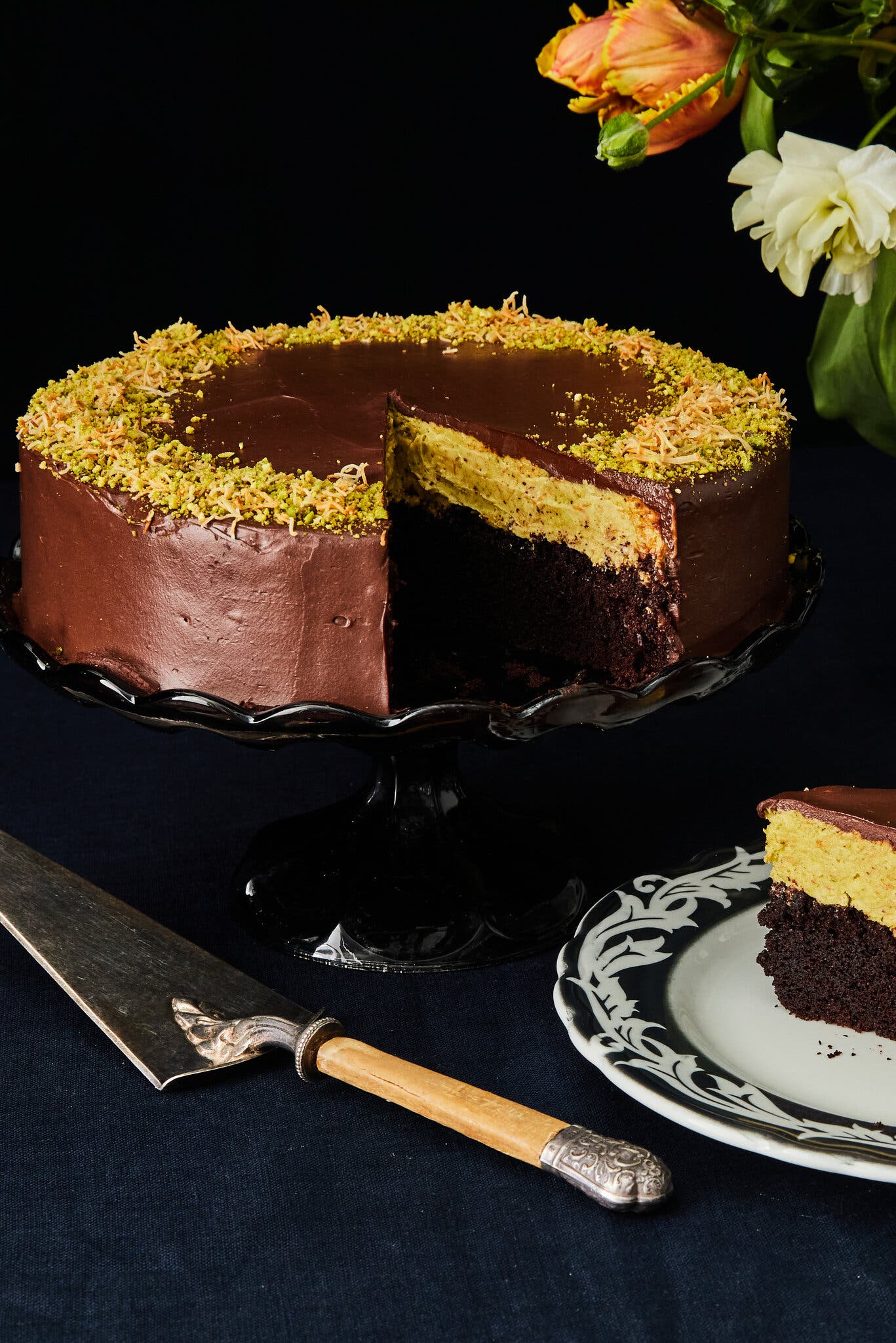 A chocolate layer cake with pistachio cream and ganache sits on a glass cake plate.