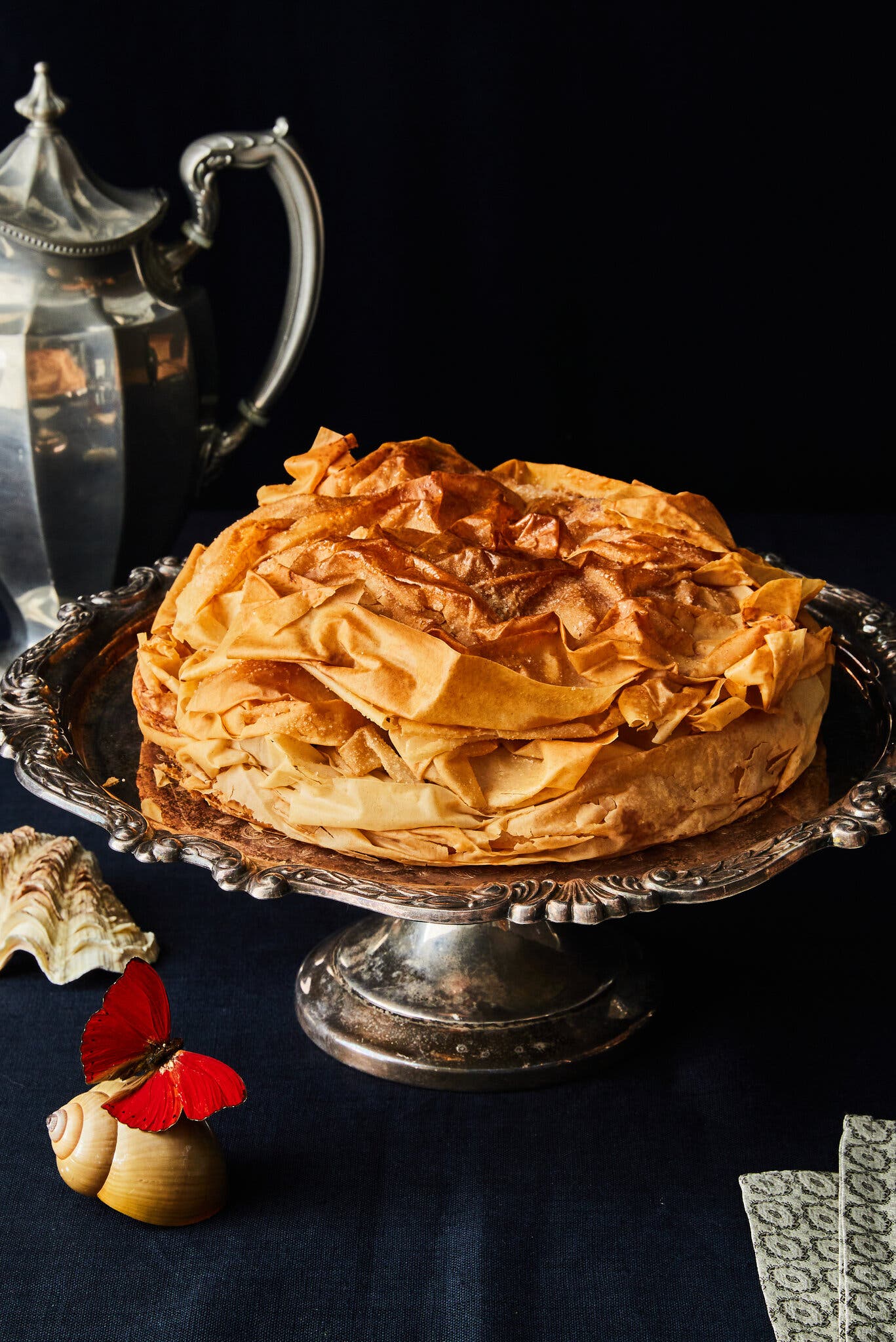 A large, golden brown phyllo pastry sits on a silver cake stand in front of a silver carafe.