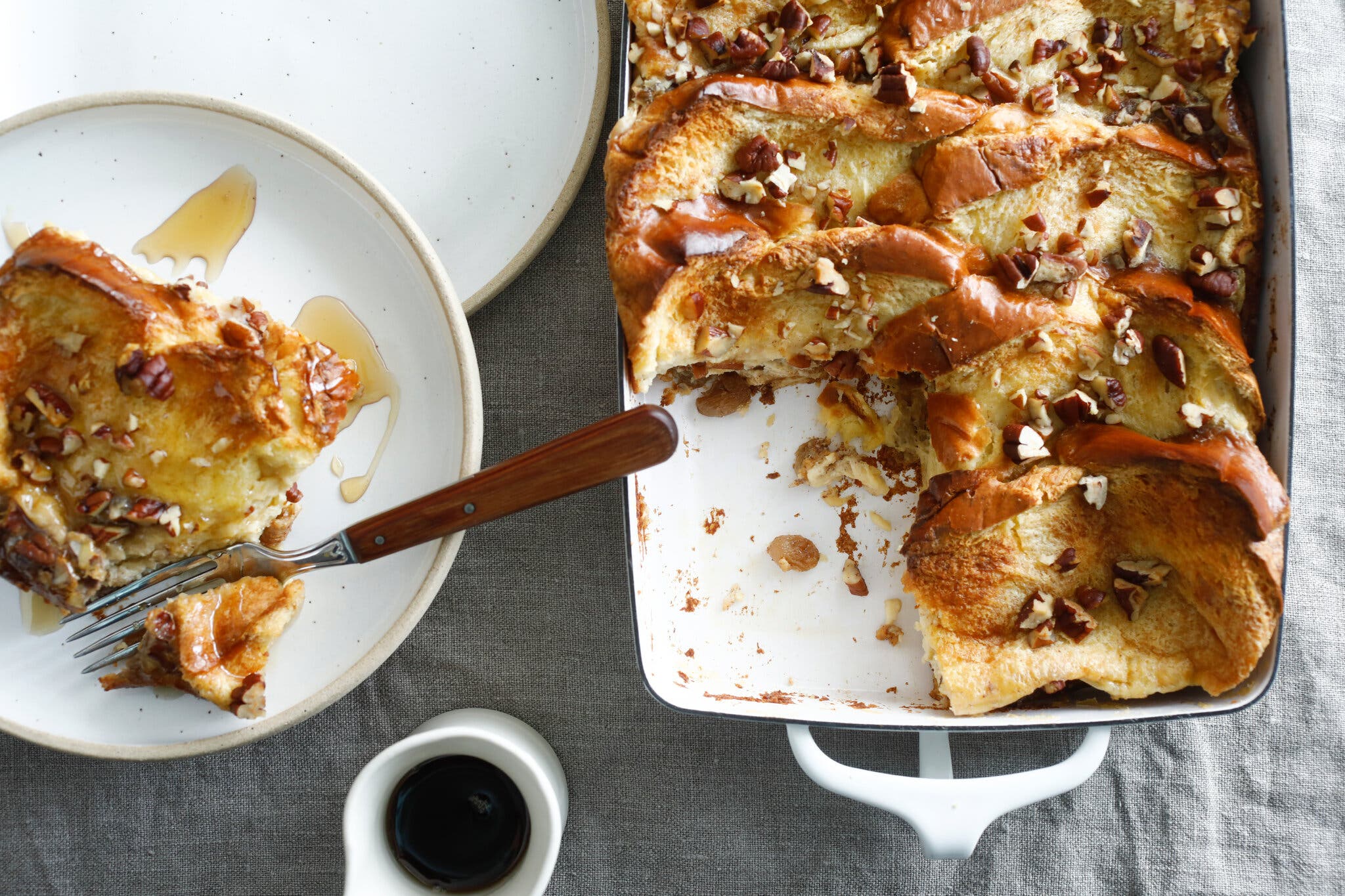 Golden-brown slices of French toast, topped with chopped nuts, fill a white baking dish. A piece on a nearby plate is drizzled with syrup.