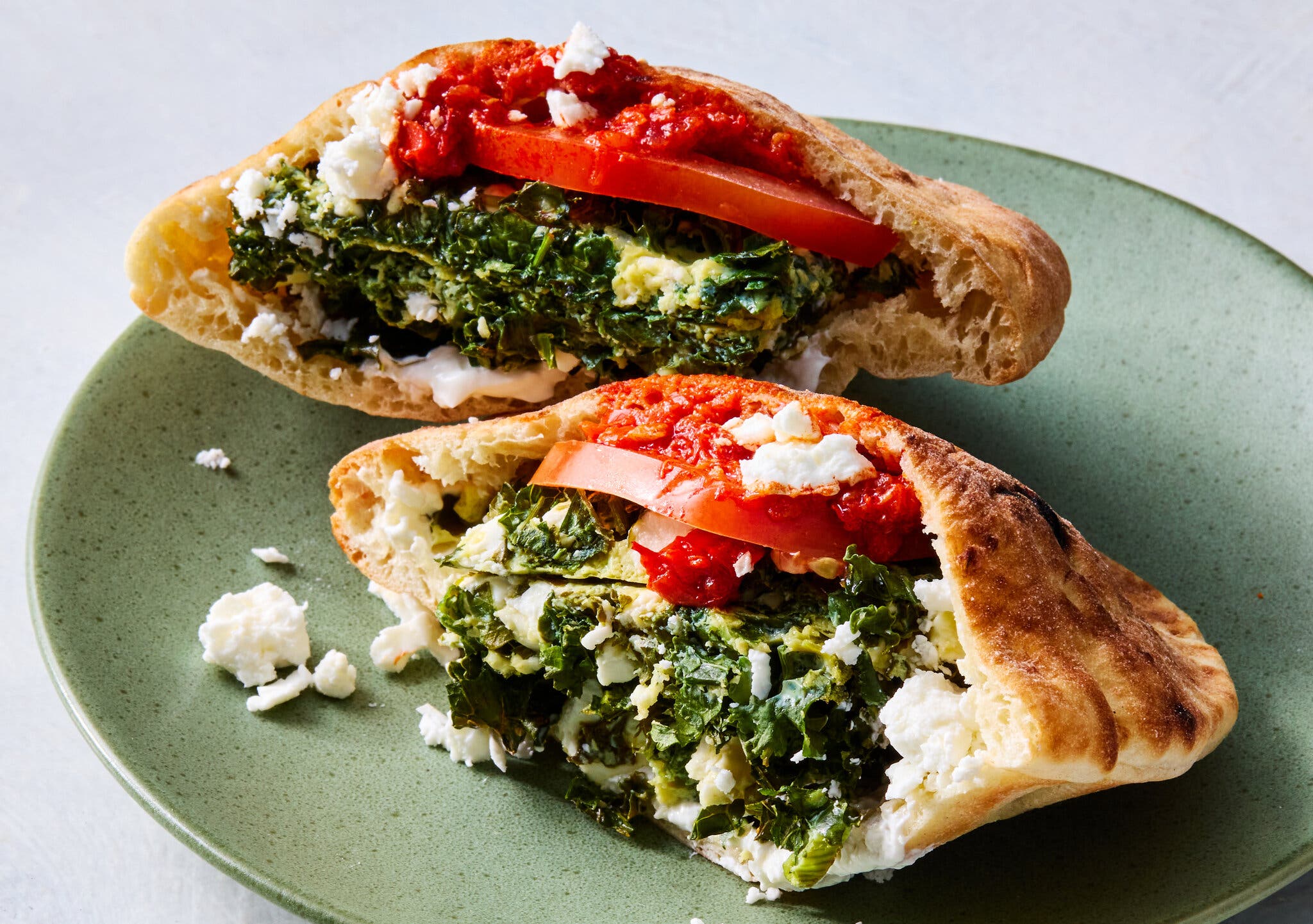 A green plate holds two open pita breads, filled with a dark green vegetable fritata, red sauce, tomato and white crumbled cheese.