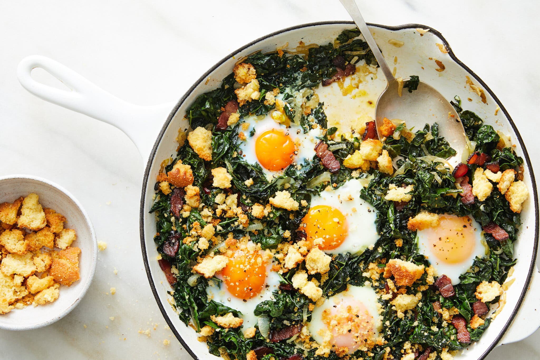 A mixture of baked eggs, greens and bits of bacon topped with golden croutons fills a white skillet. A small white bowl with more croutons sits nearby.