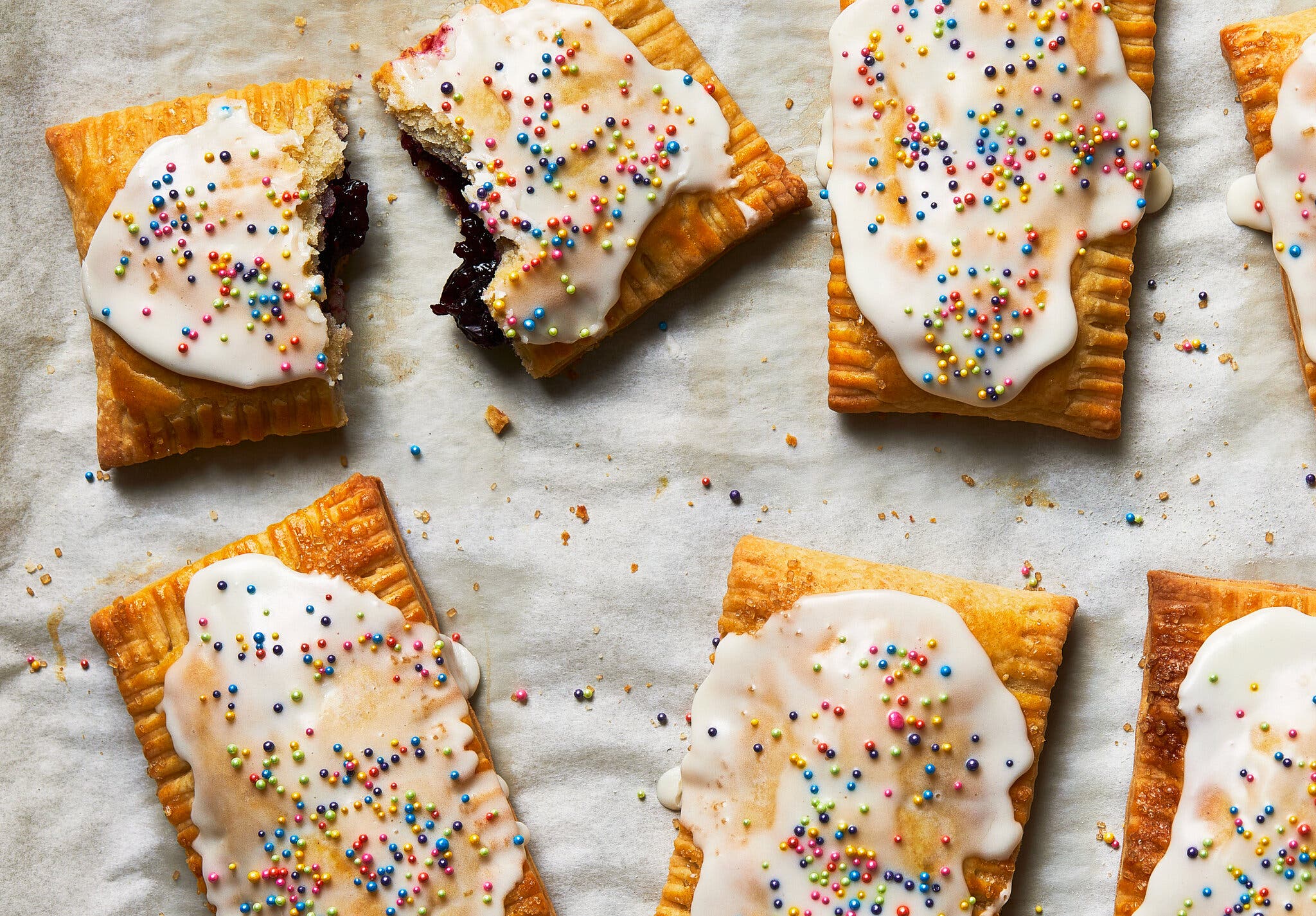 Golden rectangular pastries with white icing and colorful sprinkles on parchment paper. One pastry is broken, showing a dark filling.