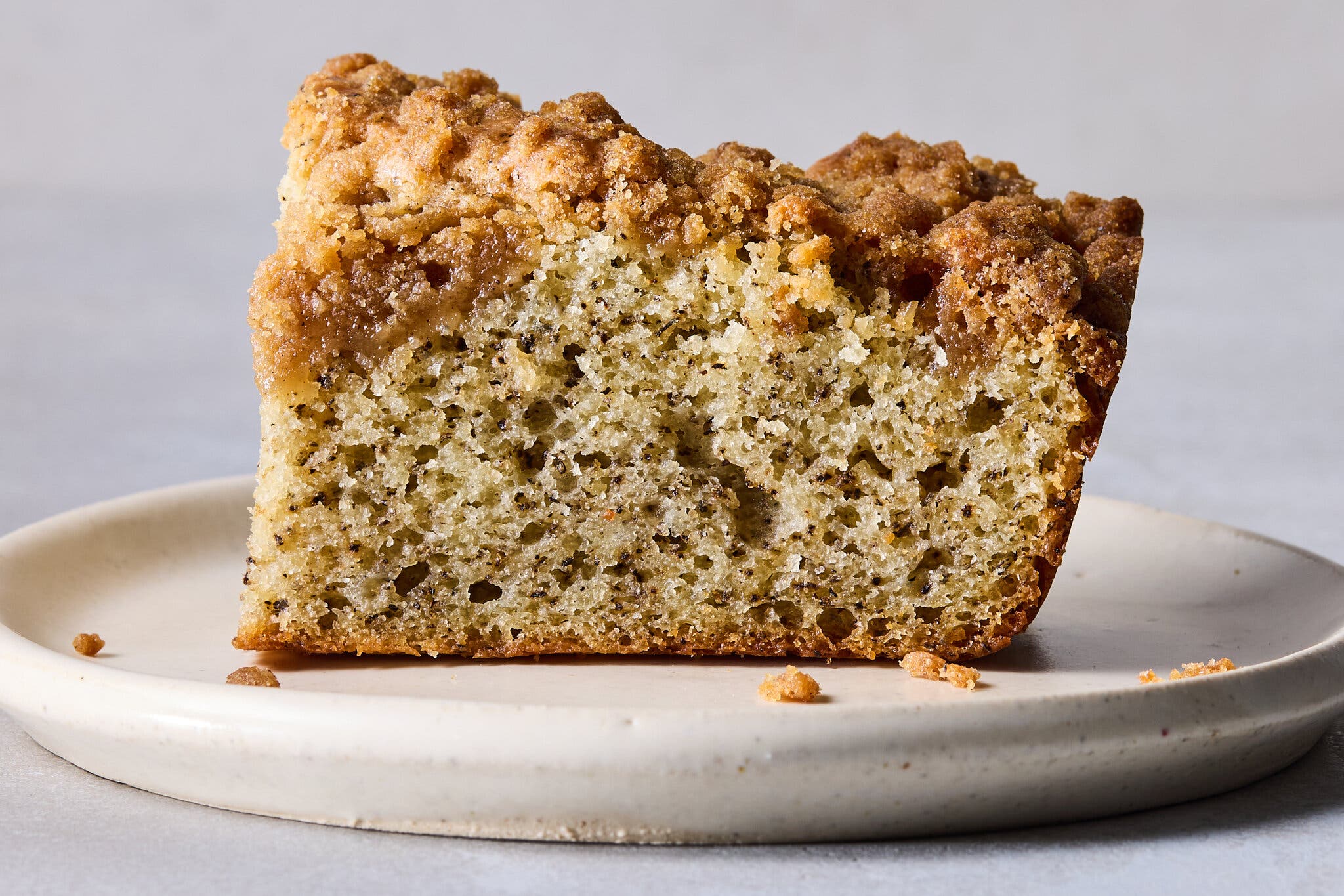 A slice of light cake with dark specks and a brown crumb topping rests on a beige plate.