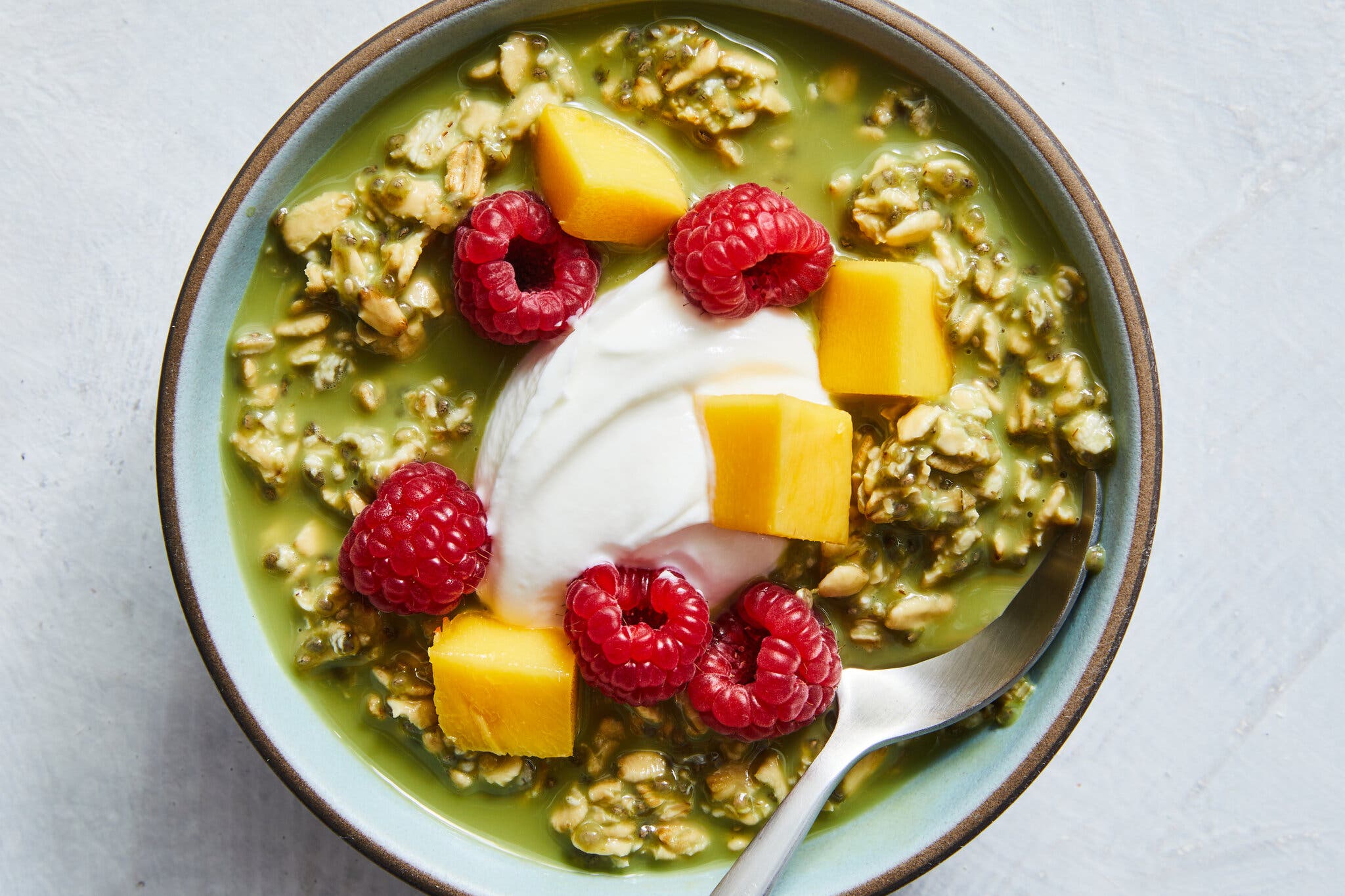 A bowl of green-tinged overnight oats topped with raspberries, cut mango and a dollop of yogurt.
