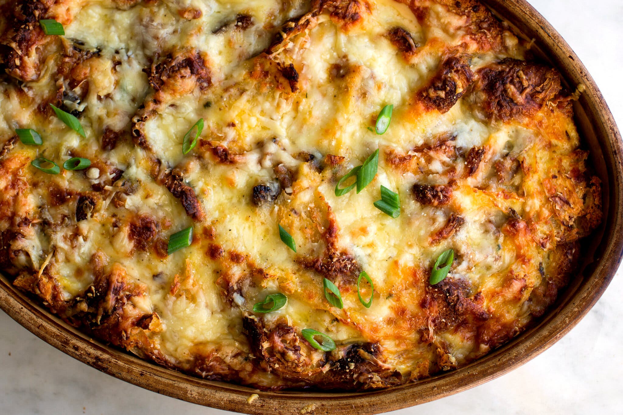 An oval, brown baking dish holds a golden-brown casserole topped with melted cheese and garnished with green scallion pieces.