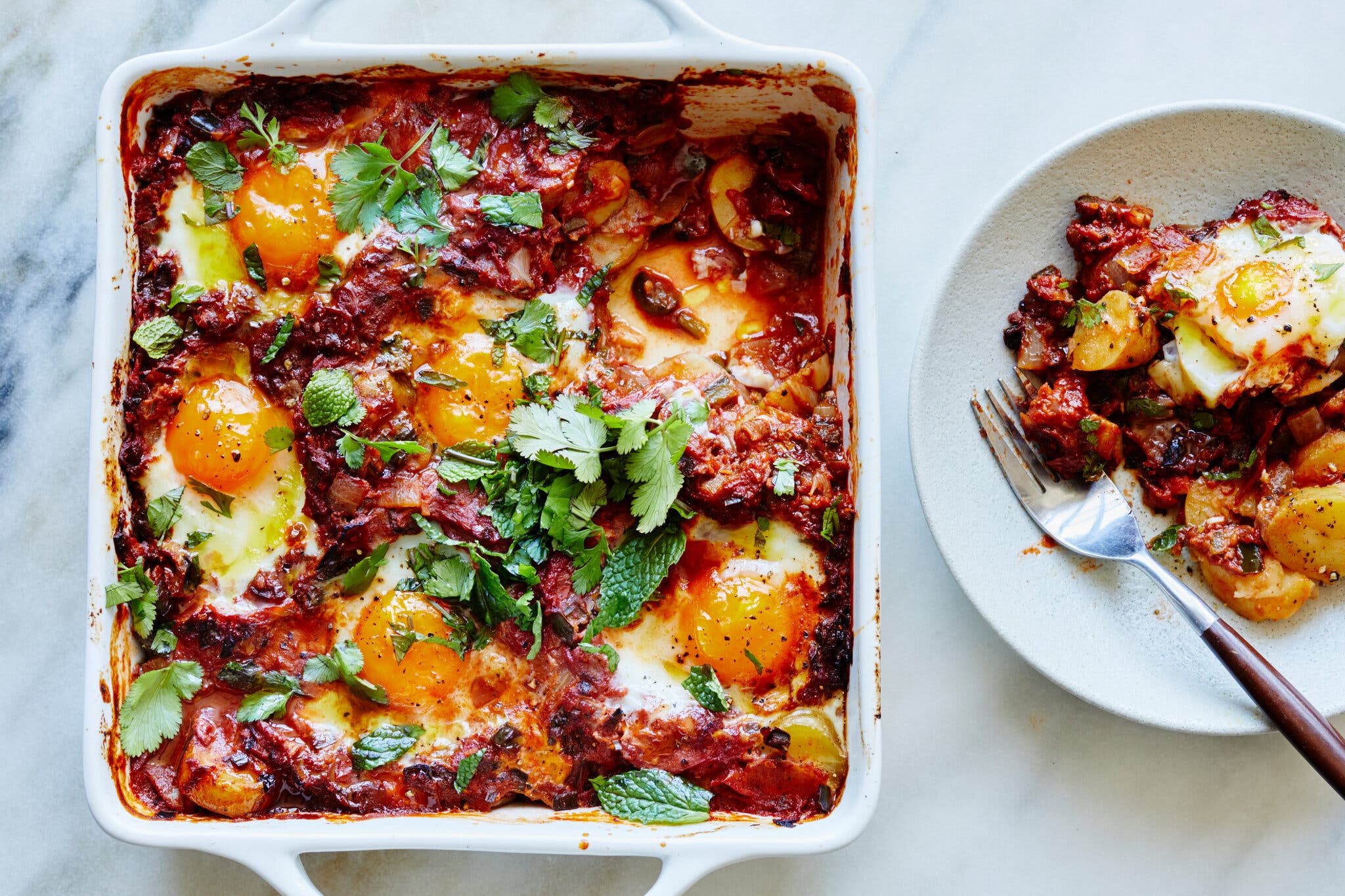 A white baking dish is filled with baked eggs and red sauce. A serving is on a white plate nearby.