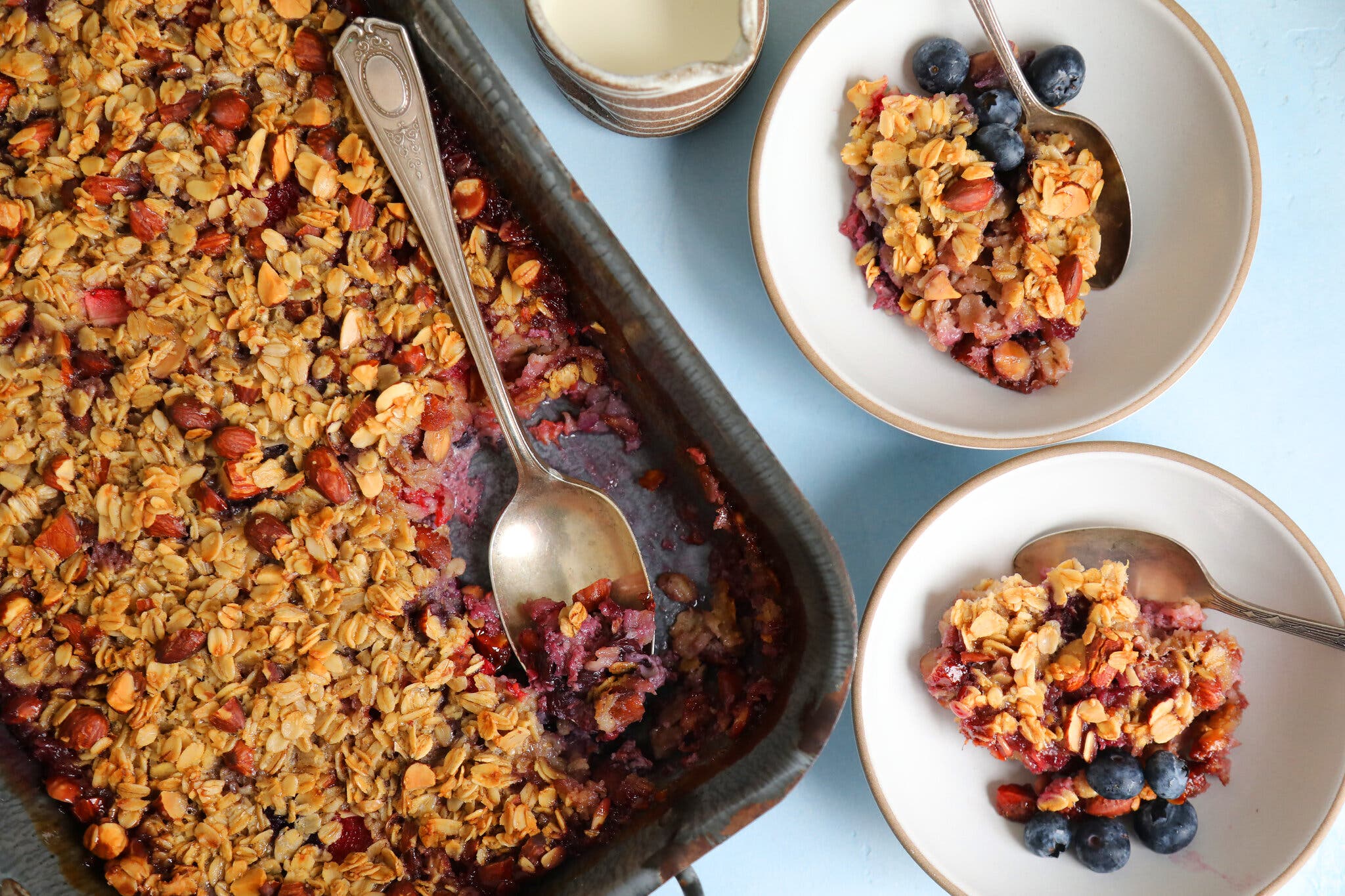 A baking dish filled with a fruit, oat and nut mixture. A silver spoon is in the dish, and two bowls of it sit nearby.