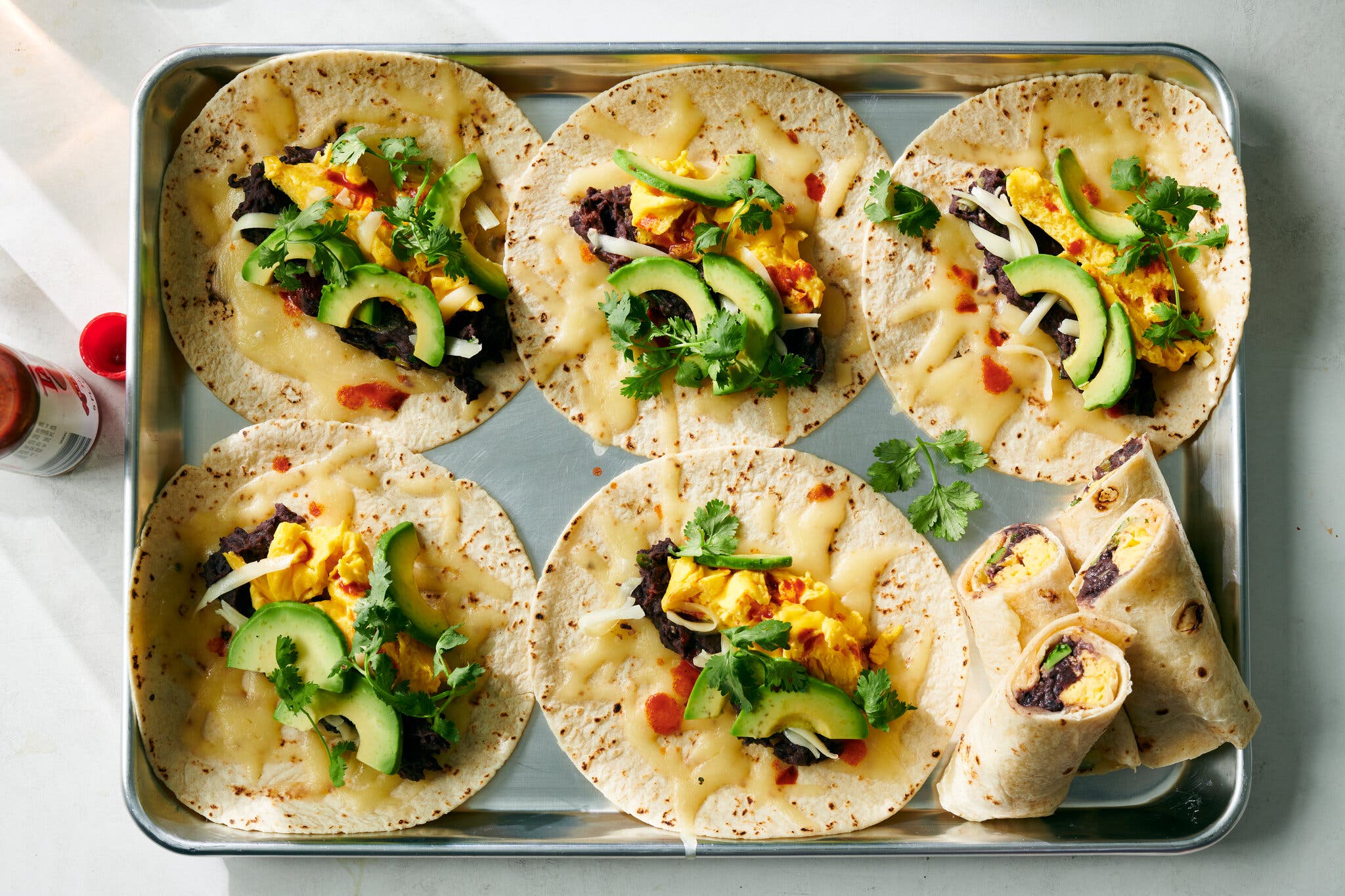 A metal tray holds tortillas topped with scrambled eggs, black beans, cheese, avocado and cilantro. Several are rolled.
