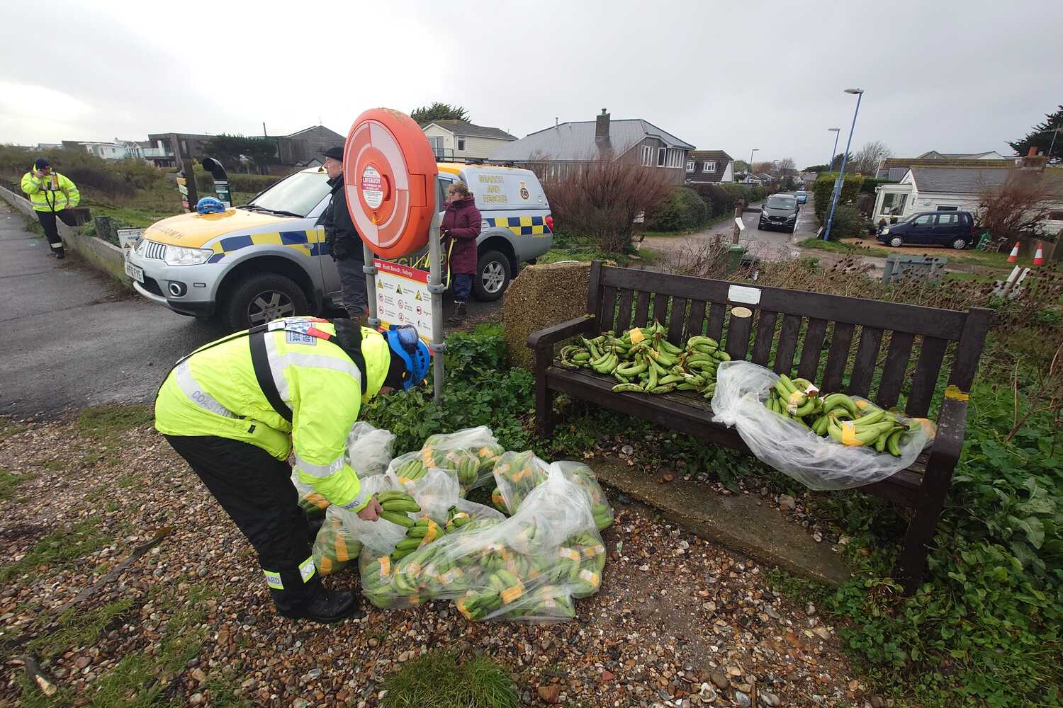 A man wearing a high-viz yellow coat standing in front of several plastic bags full of green bananas. Even more bananas sit on a bench in front of him.