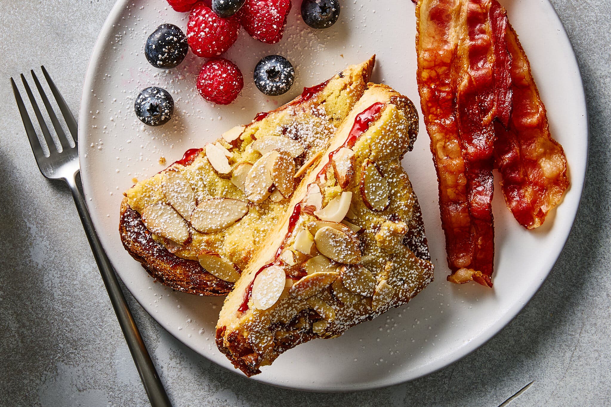 Two wedges of French toast stuffed with jam, topped with sliced almonds and powdered sugar, served with crispy bacon and fresh berries.