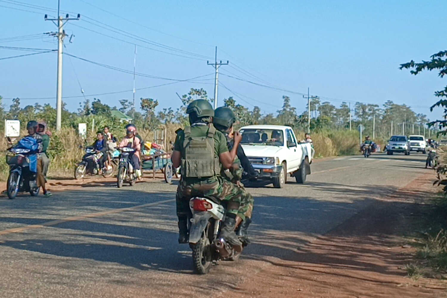Two people dressed in military fatigues on a motorbike. They are on a small road, where almost all of the other traffic is moving in the opposite direction.