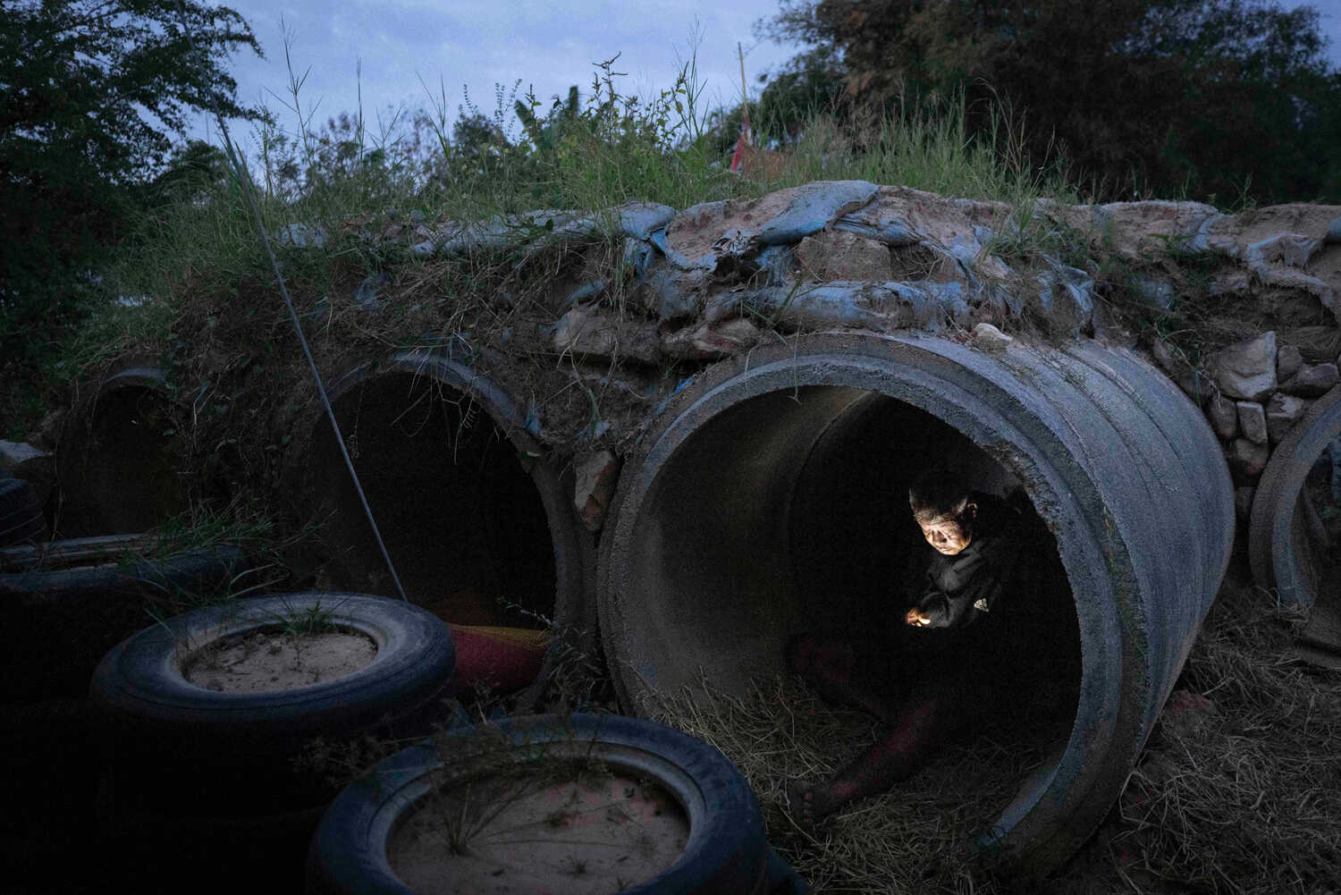 A person, whose face is illuminated by a phone, sits in a dark, empty pipe, surrounded by old tires and rocks.