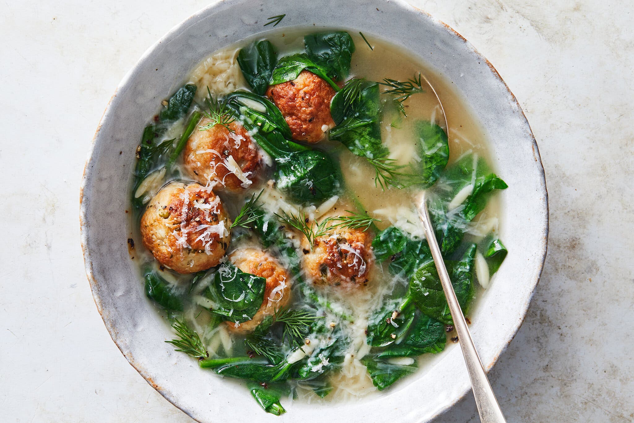 An overhead image of a bowl of Italian wedding soup with large meatballs, Parmesan cheese, orzo and spinach, garnished with dill.