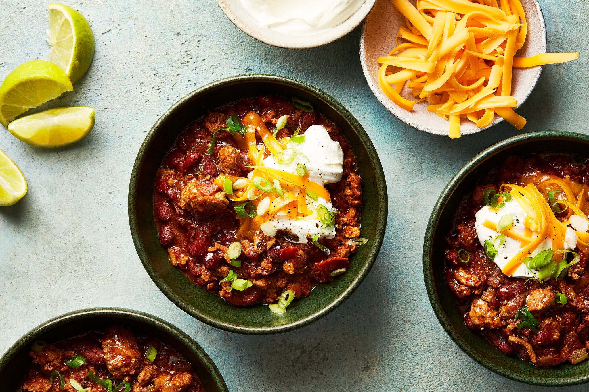 An overhead image of several bowls of turkey chili, garnished with sour cream, scallions and Cheddar. Nearby are lime wedges, a bowl of sour cream and a bowl of more shredded Cheddar.