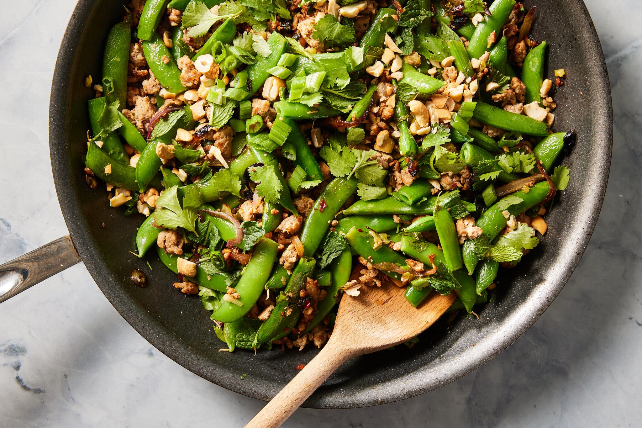An overhead image of a skillet of snap peas and ground turkey with sliced scallions and ginger. There is a wooden spoon in the skillet.