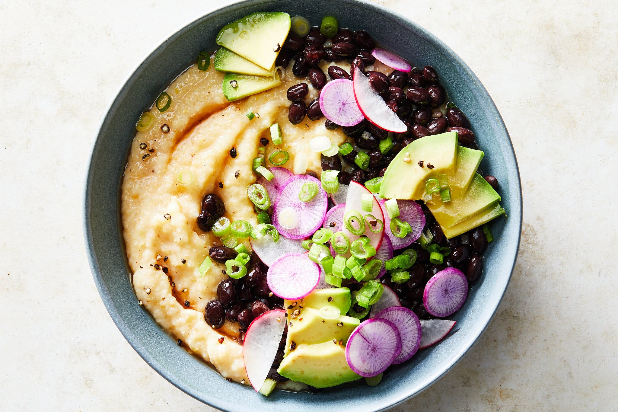 A blue bowl holds cheese grits with black beans, avocado, radishes and scallions.