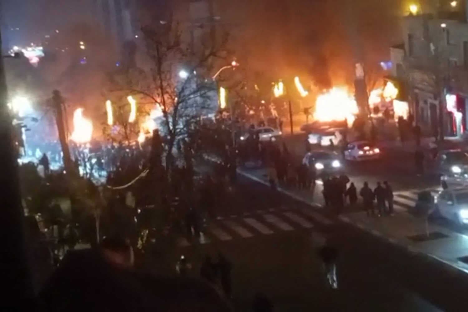 A blurry image of a dark street filled with fires, cars and people.